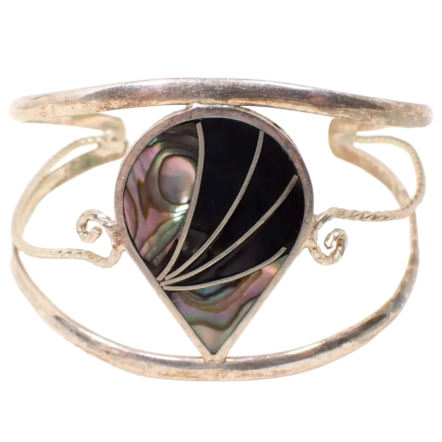 Enlarged front view of the Alpaca Mexican bracelet. The metal is silver tone in color. There are three rounded bands with open spaces between them making up the cuff part of the bracelet. The top and bottom are curved. The middle one has a twisted design with curls at the end. In the middle is a teardrop shape with inlaid abalone shell and black enamel with silver tone color metal curves between the areas.
