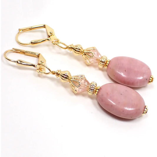 Angled view of the handmade rhodonite earrings. The metal is gold plated in color. There are faceted glass light pink crystal beads at the top. The gemstones are puffy oval shaped and have shades of soft rosy pink.