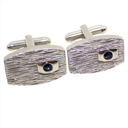 Enlarged angled view of the Mid Century vintage Modernist cufflinks. They are rectangle like shapes with curved out bottom and top sides. There is a line textured front with small a small dark blue rhinestone on one side. On one of the cufflinks you can see a small area where the silver tone plating has been chipped over time.