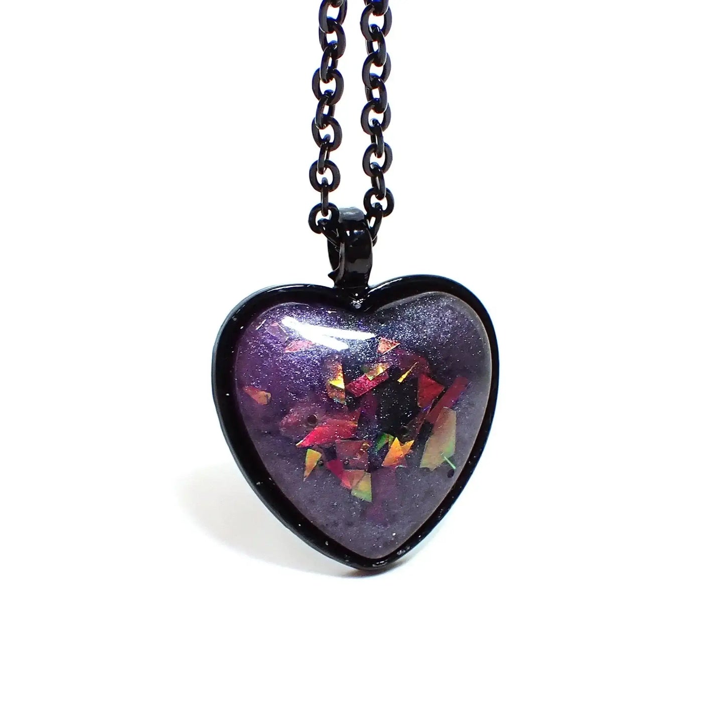 Enlarged front view of the Goth black heart pendant necklace. The chain and setting are black coated. There is a domed heart pendant at the bottom with a handmade resin cab. The resin has pearly shades of purple resin. There are pieces of chunky iridescent glitter embedded in it that show different colors depending on how the light hits it.