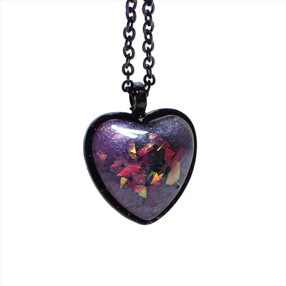 Enlarged front view of the Goth black heart pendant necklace. The chain and setting are black coated. There is a domed heart pendant at the bottom with a handmade resin cab. The resin has pearly shades of purple resin. There are pieces of chunky iridescent glitter embedded in it that show different colors depending on how the light hits it.