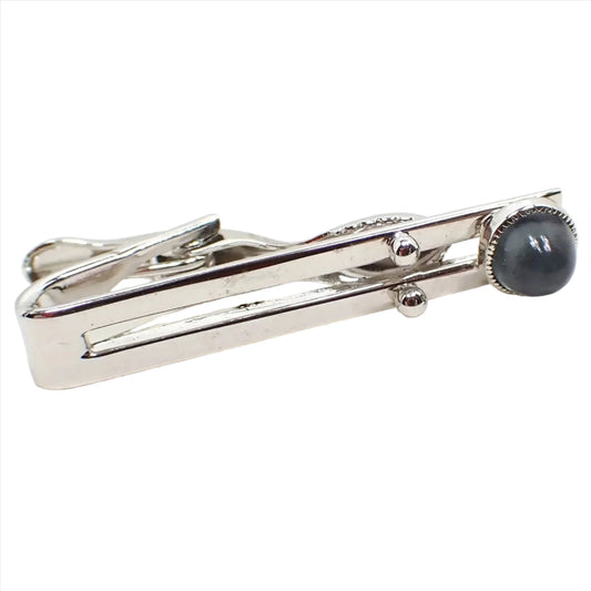 Front view of the retro vintage lucite tie clip. The metal is silver tone in color. There front bar has an open middle with two round balls towards the end. The very end has a round high domed small lucite cab in a bluish gray color.