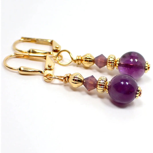 Angled view of the handmade amethyst earrings. The metal is gold tone in color. There are small purple glass crystal faceted beads at the top. The gemstone beads on the bottom are round sphere shaped and are a darker shade of purple in color.