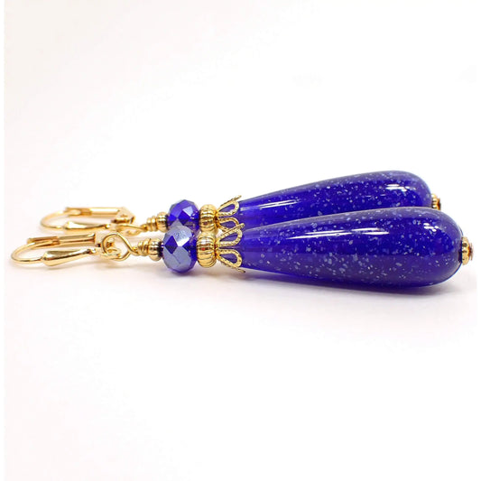 Side view of the handmade teardrop earrings. The metal is gold plated in color. There is a faceted cobalt blue glass crystal bead at the top. The bottom bead is a rounded teardrop shape with dark blue lucite and small flecks of white confetti. The confetti lucite beads are vintage beads that were never before used.