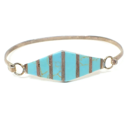 Front view of the Alpaca retro vintage Mexican bangle bracelet. The metal is silver tone in color and has a slightly darkened patina from age. The front has a diamond shape with metal stripes. In between the stripes are inlaid pieces of blue resin with hints of brown to look like turquoise.