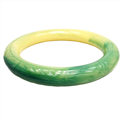 Angled view of the Mid Century vintage lucite bangle bracelet. Half of the bangle is green in color and the other is yellow and they blend together where the two meet. There is a lightly carved left design around the outer edge.