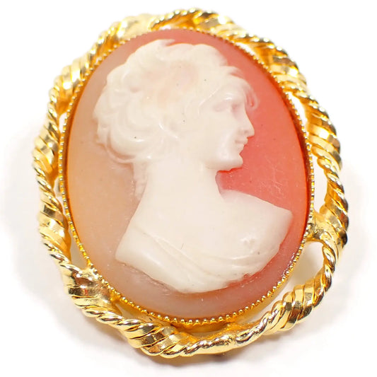 Enlarged front view of the retro vintage molded plastic cameo brooch. The setting is gold tone and has a twisted design around the edge. It is oval in shape with a bust of a woman on the front. The background goes from light peach to a salmon pink in color.