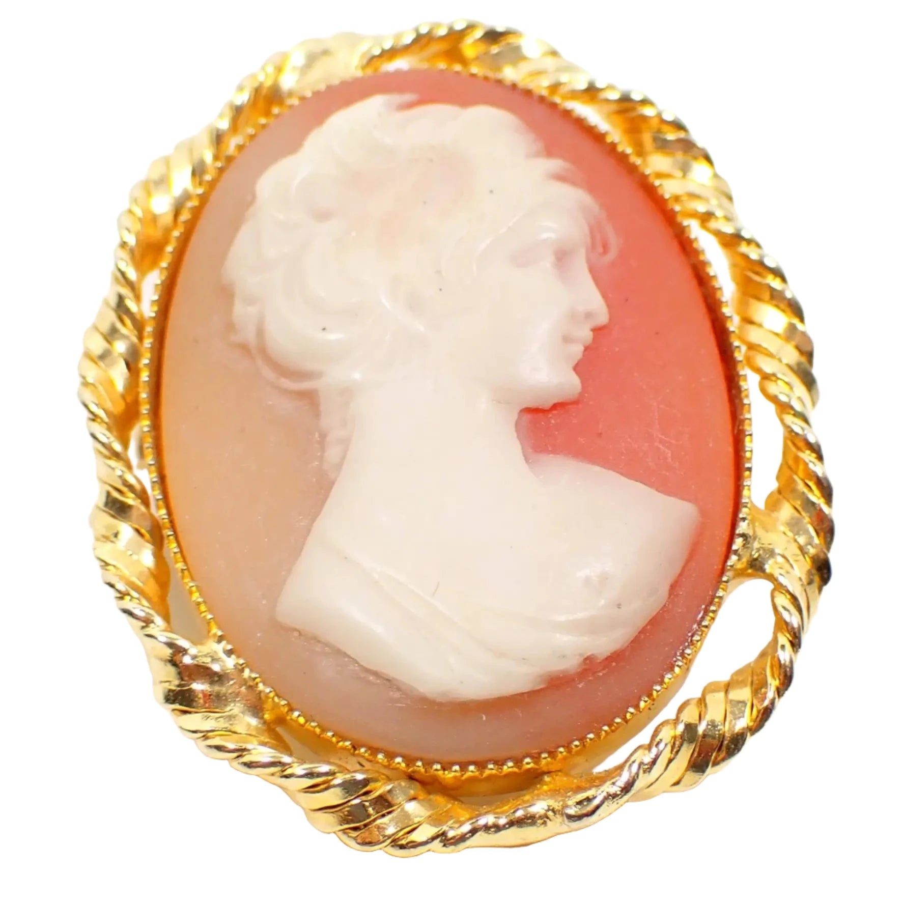 Enlarged front view of the retro vintage molded plastic cameo brooch. The setting is gold tone and has a twisted design around the edge. It is oval in shape with a bust of a woman on the front. The background goes from light peach to a salmon pink in color.
