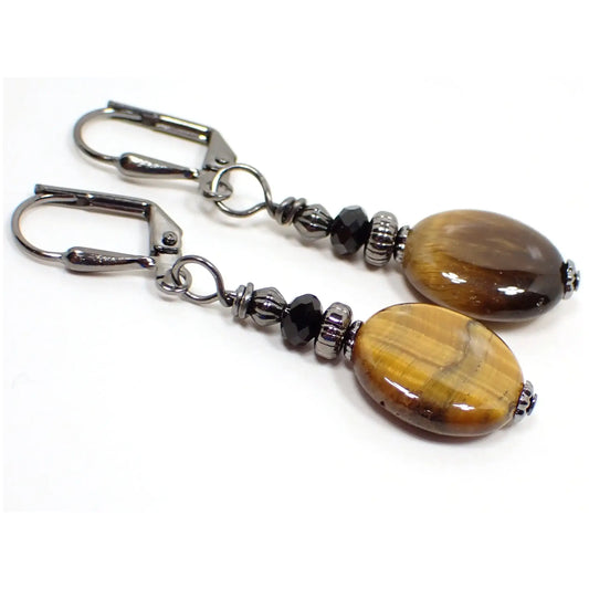 Angled view of the handmade gemstone earrings. The metal is gunmetal gray in color. There are faceted black glass crystal beads at the top. The bottom beads are oval tiger's eye gemstone and have shades of brown and yellow that have a shimmer effect as you move around in the light.
