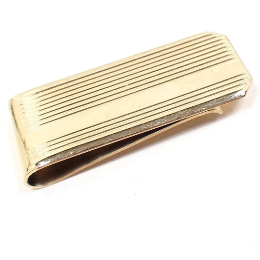 Angled top view of the retro vintage money clip. The metal is gold tone in color. It is rectangular shaped with a faceted end. There are thin engraved stiped lines on the front with a wider stripe towards the bottom.