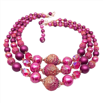 Front view of the Mid Century vintage multi strand beaded necklace from Japan. There are three strands of beads with gold tone plated bead caps at the bottom and hook clasp at the end. The top part of the necklace has round bright pink pearly beads and matte fuchsia purple beads. Going down the sides and bottom part of the necklace are bumpy textured pink beads with metallic gold highlights, pink beads with some splashes of metallic gold and faceted AB red oval beads.