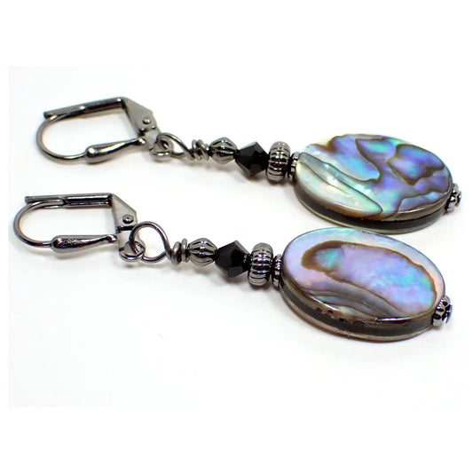 Angled view of the handmade abalone earrings. The metal is gunmetal gray in color. There is a black faceted glass crystal bead at the top. The bottom abalone shell beads are flat oval shaped and have shades of gray, purple, blue, and green that have some flash as you move around in the light.