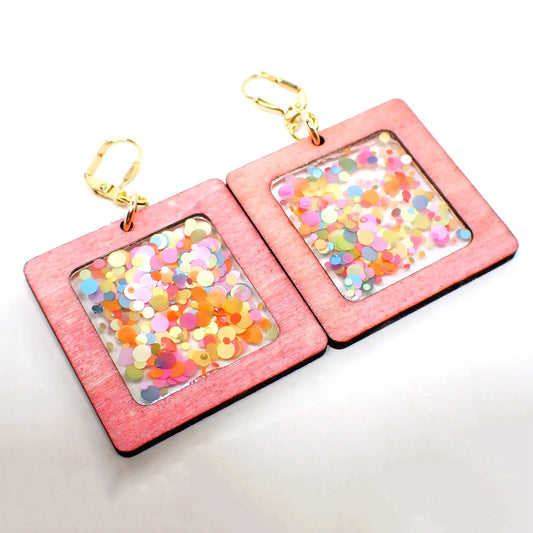 Angled front view of the handmade geometric square drop earrings. The metal is gold tone plated in color. The drops are large square shapes with pink dyed wood. The middle has clear resin with pastel rainbow color round confetti dots.