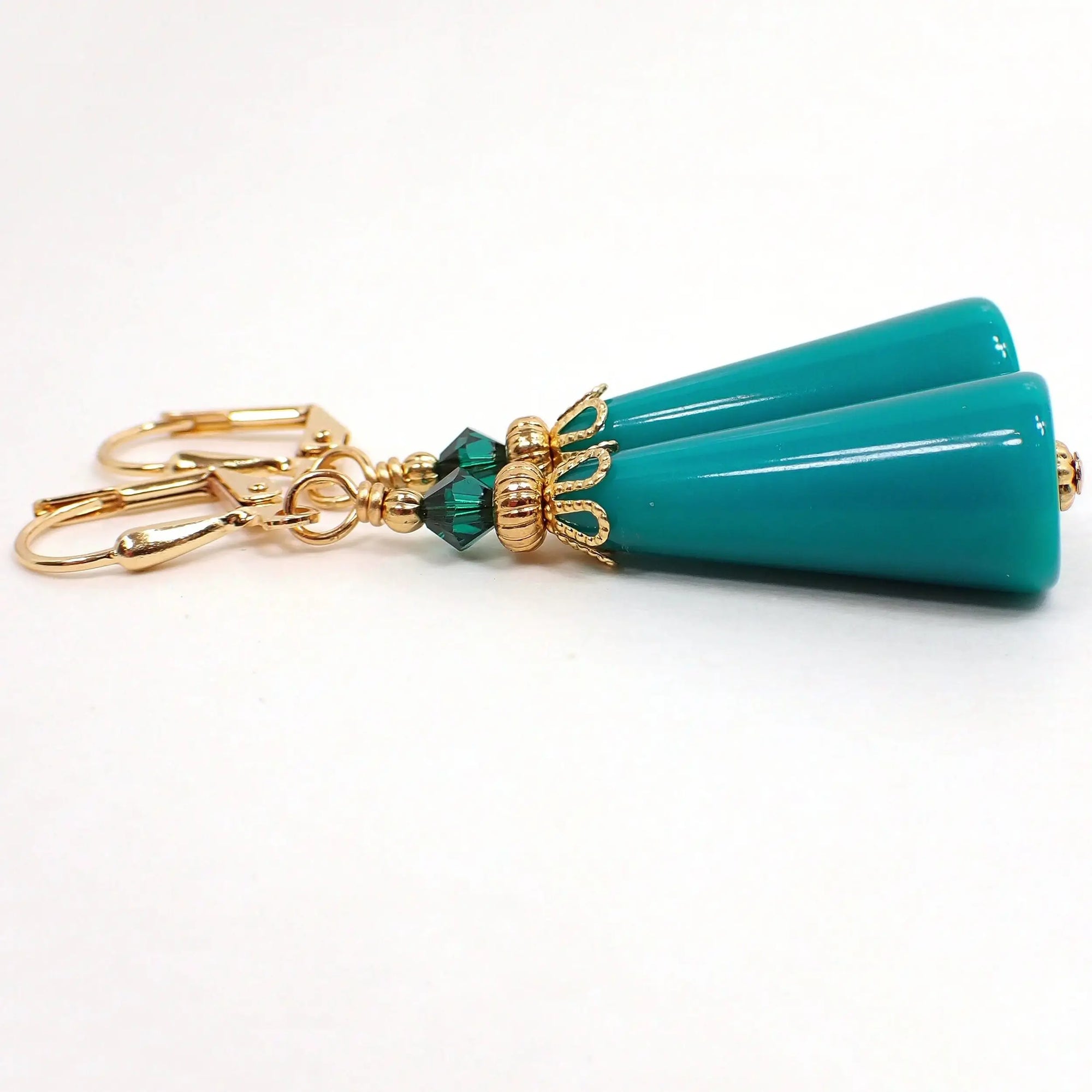 Side view of the handmade geometric cone earrings. The metal is gold plated in color. There is a faceted dark green glass crystal bead at the top. The bottom cone shaped bead is a vintage lucite bead in teal green.