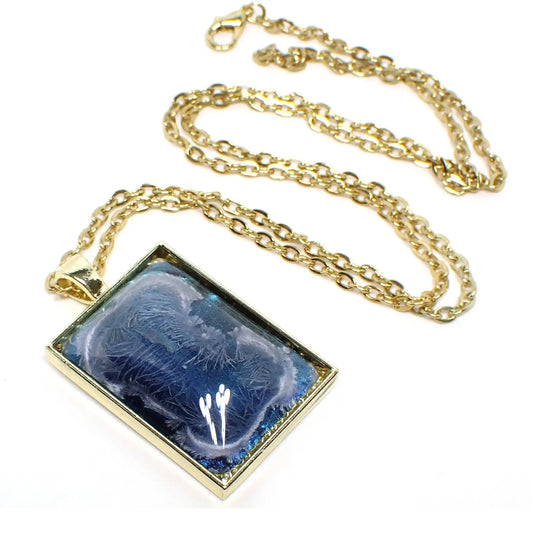 Angled front view of the large handmade rectangle pendant necklace. The metal is gold tone plated in color. There is a domed resin cab that has an iridescent blue background with hints of green. There is an abstract frost like design in a misty white color.