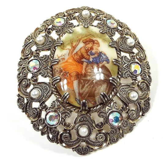 Front view of the Mid Century vintage West Germany cameo brooch pin. It is oval in shape and has a filigree light gold tone metal design. There is a prong set painted cameo cab in the middle depicting two people sitting under a tree holding flowers. There are AB rhinestones and faux pearls around the edge of the brooch.