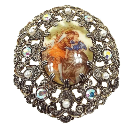 Front view of the Mid Century vintage West Germany cameo brooch pin. It is oval in shape and has a filigree light gold tone metal design. There is a prong set painted cameo cab in the middle depicting two people sitting under a tree holding flowers. There are AB rhinestones and faux pearls around the edge of the brooch.