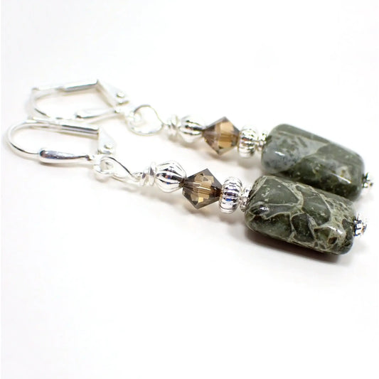 Angled view of the handmade green rhyolite gemstone earrings. The metal is silver plated in color. There are smoky brown faceted glass crystal beads at the top. The bottom gemstone beads are puffy square in shape and have a chunky pattern with shades of green, cream, and hints of brown.
