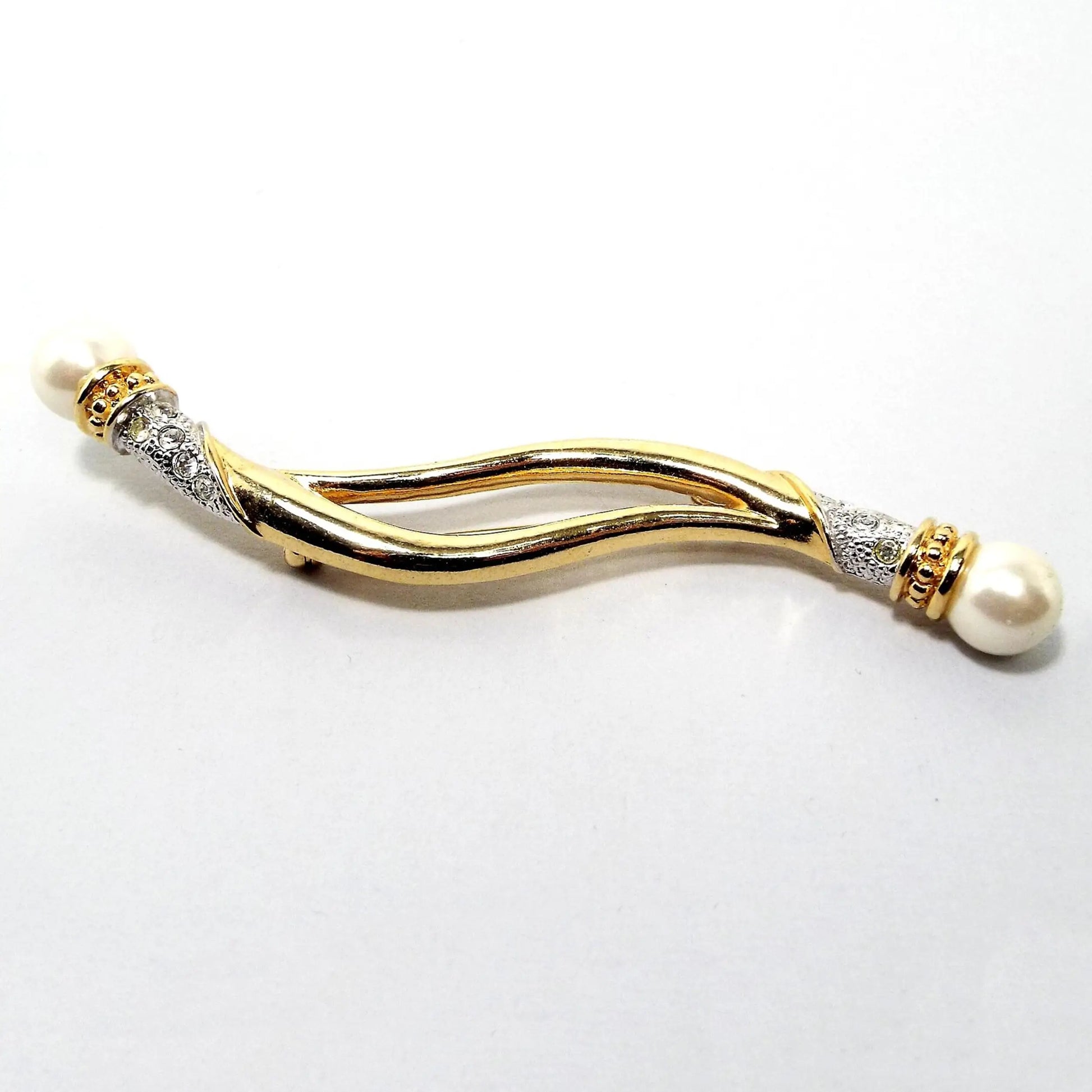 Front view of the retro vintage Avon brooch pin. It is long and curvy with an open split area in the middle. The metal is gold tone in color except for at the ends where it's silver tone in color. In the silver tone area are small round clear rhinestones. There are round faux pearls at the ends.