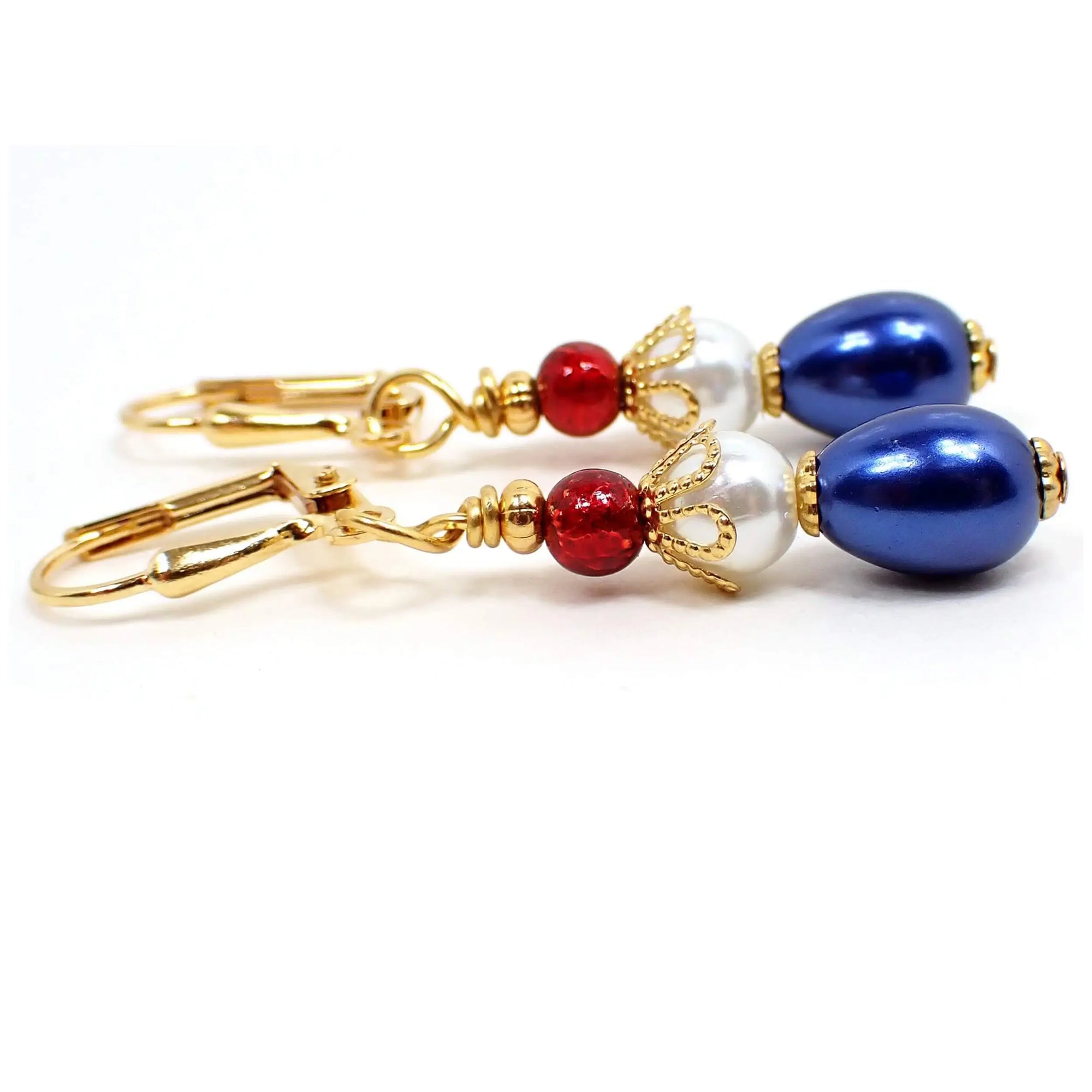 Enlarged side view of the red white and blue handmade earrings. The metal is gold plated in color. There is a small metallic red glass bead at the top, a white faux pearl bead in the middle, and a metallic blue teardrop bead at the bottom.