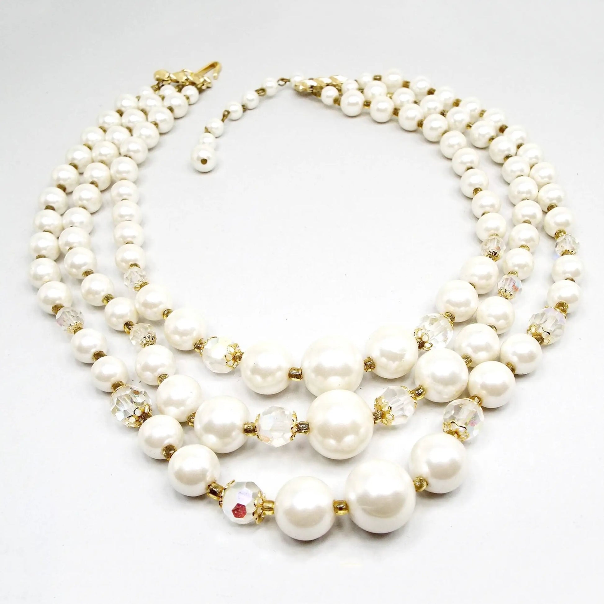 Front view of the Japanese Mid Century vintage multi strand necklace with AB crystal and faux pearl beads. There are three beaded strands with a gold tone color hook clasp at the end. Each strand has white plastic imitation pearl beads and a few AB glass crystal beads towards the bottom of the necklace. There are small gold tone color beads in between the other beads as spacers.