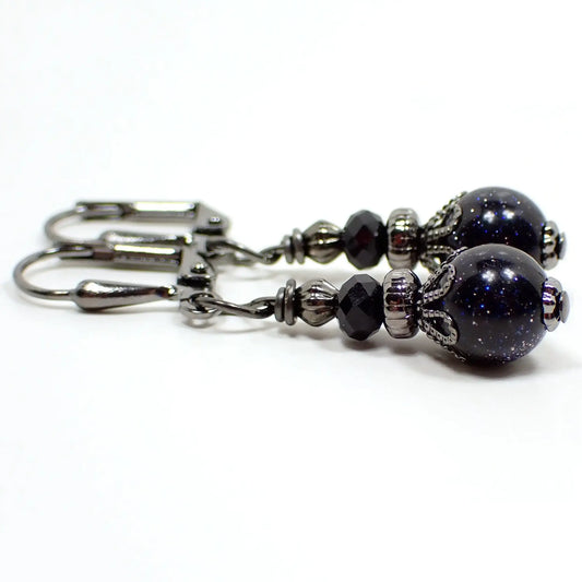 Photo of the small handmade blue goldstone drop earrings. The metal is gunmetal gray in color. There is a small faceted black glass crystal bead at the top. The bottom bead is round ball shaped and is blue goldstone, which is a dark blue glass with fine and tiny blue and silver flecks of metallic glitter.
