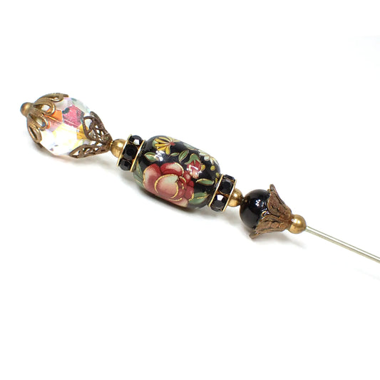 Enlarged angled view of the top of the retro vintage stick pin. The top has a faceted AB crystal glass bead with filigree cap on top and bottom. There is a plastic barrel bead below that which has a floral decal design with a black background and black rhinestone beads above and below it. The very bottom bead is round black glass with a filigree cap on the bottom. The metal is antiqued brass in color.