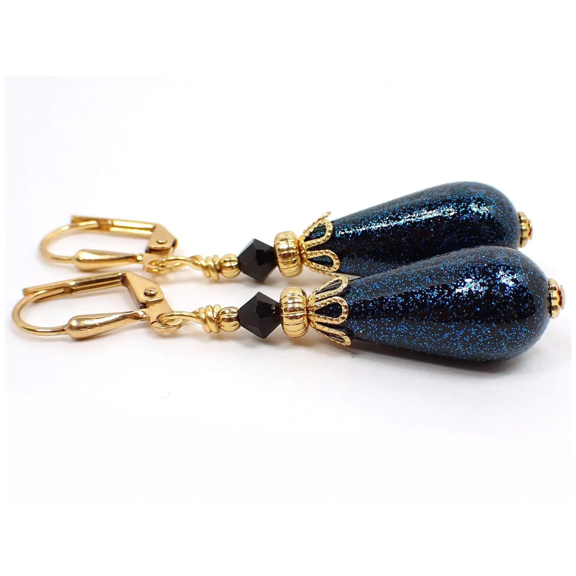 Side view of the handmade teardrop earrings with vintage German acrylic beads. The metal is gold plated in color. There are faceted black glass crystals at the top. The bottom acrylic beads are teardrop shaped and are black with tiny flecks of teal blue glitter embedded in them for sparkle all the way around.