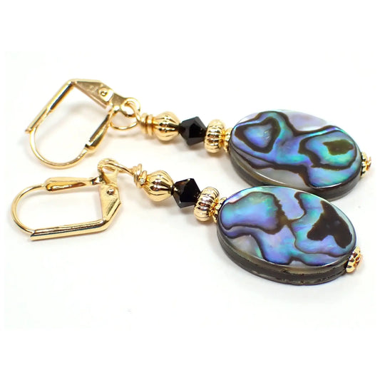 Photo of the handmade abalone earrings. The metal is gold plated in color. There is a black faceted glass crystal bead at the top. The bottom abalone shell beads are flat oval shaped and have shades of gray, purple, blue, and green that have some flash as you move around in the light.