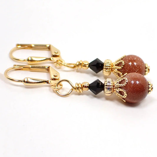 Side view of the handmade goldstone drop earrings. The metal is gold plated in color. There are faceted black crystal glass bicone beads at the top and small round ball goldstone beads at the bottom. The goldstone beads are orange glass with tiny flecks of copper for sparkle.