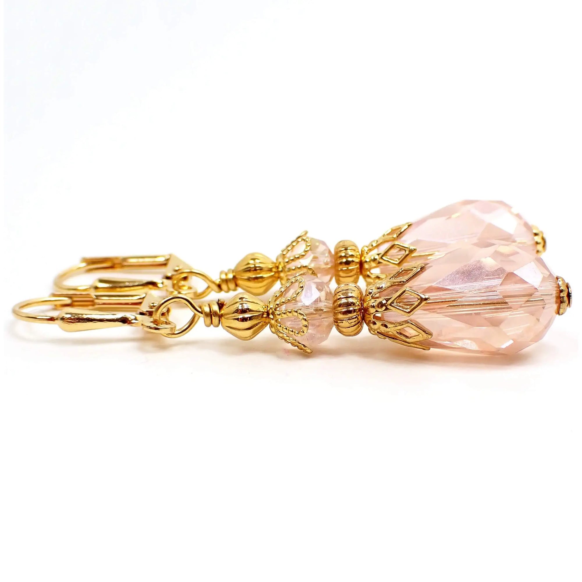 Side view of the light pink teardrop earrings. The metal is gold plated in color. There are faceted glass crystal rondelle beads at the top and faceted glass crystal teardrop beads at the bottom. The beads are a light pink in color.