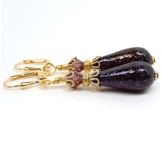 Side view of the handmade teardrop earrings with vintage German acrylic beads. The metal is gold plated in color. There are faceted purple glass crystals at the top. The bottom acrylic beads are teardrop shaped and are black with tiny flecks of purple glitter embedded in them for sparkle all the way around.
