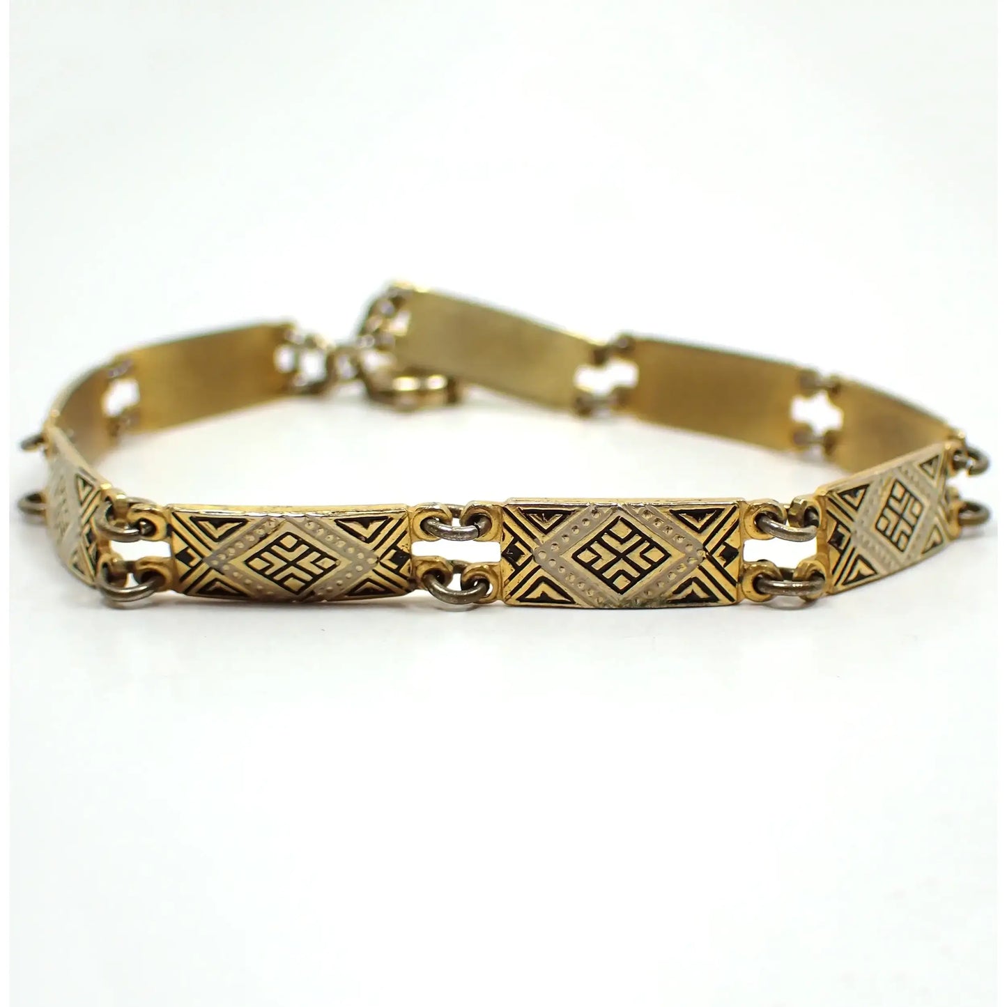 Enlarged view of the the bracelet on its side showing the top side of some of the links. They are rectangle shaped and the metal is gold tone plated in color. There is a geometric diamond shaped design in the middle with a white painted edge and black painted accents on the rest of the link. The jump rings holding the links together are darkened. There is a spring ring clasp showing at the back of the photo.