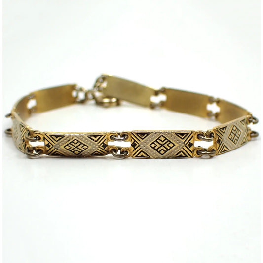Enlarged view of the the bracelet on its side showing the top side of some of the links. They are rectangle shaped and the metal is gold tone plated in color. There is a geometric diamond shaped design in the middle with a white painted edge and black painted accents on the rest of the link. The jump rings holding the links together are darkened. There is a spring ring clasp showing at the back of the photo.