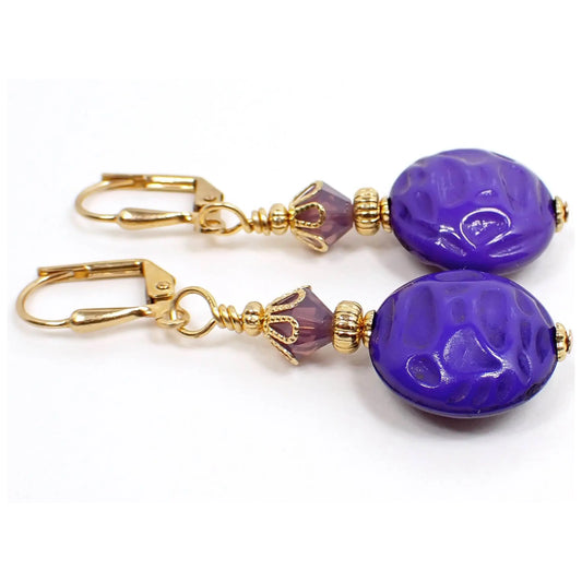 Angled view of the handmade drop earrings. The metal is gold plated in color. There are faceted light purple crystal glass beads at the top. The bottom vintage lucite beads are round circle shaped and have a bumpy indented textured pattern. They are a bright purple color that takes on shades of blue depending upon what lighting you're in.