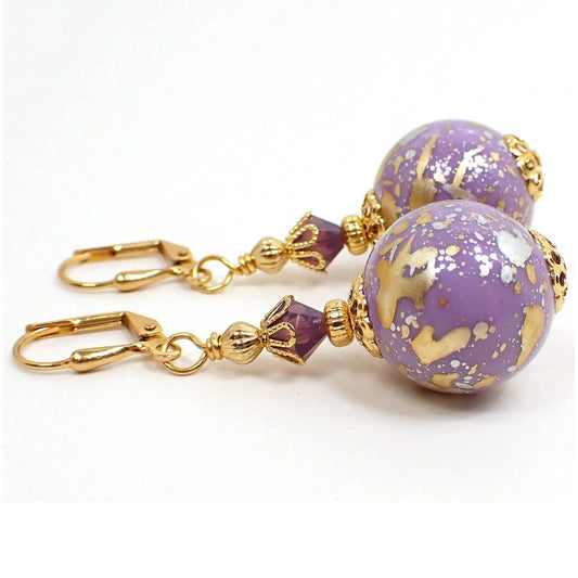 Side view of the handmade acrylic ball bead drop earrings. The metal is gold plated in color. There is a faceted glass crystal bead in a light purple at the top. The bottom bead is round sphere shaped and light purple in color. There is a metallic splash design on the bottom beads with metallic gold and silver paint.