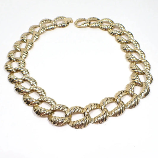 Angled top view of the retro vintage chain necklace. The metal is gold tone plated in color. There are large oval curb links with a diagonal line corrugated pattern. There is a snap lock clasp at the end.