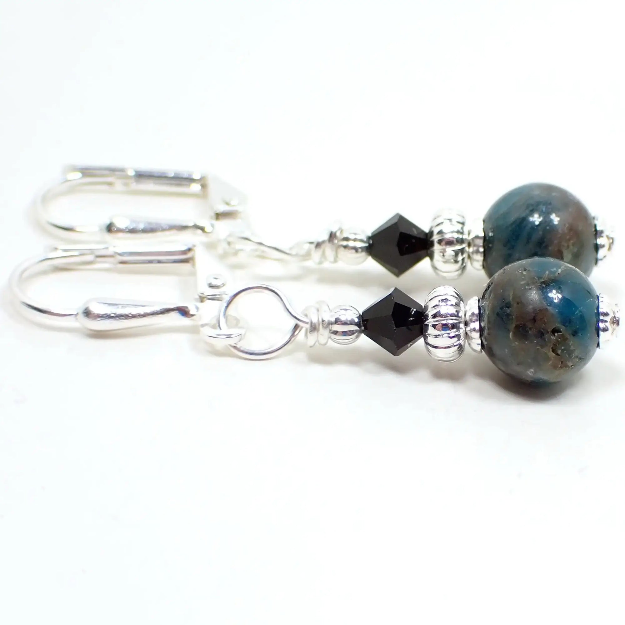 Enlarged side view of the small handmade apatite drop earrings. The metal is silver plated in color. There is a faceted glass crystal rondelle bead at the top and a small round ball apatite gemstone bead at the bottom. The gemstone is a marbled teal blue in color.