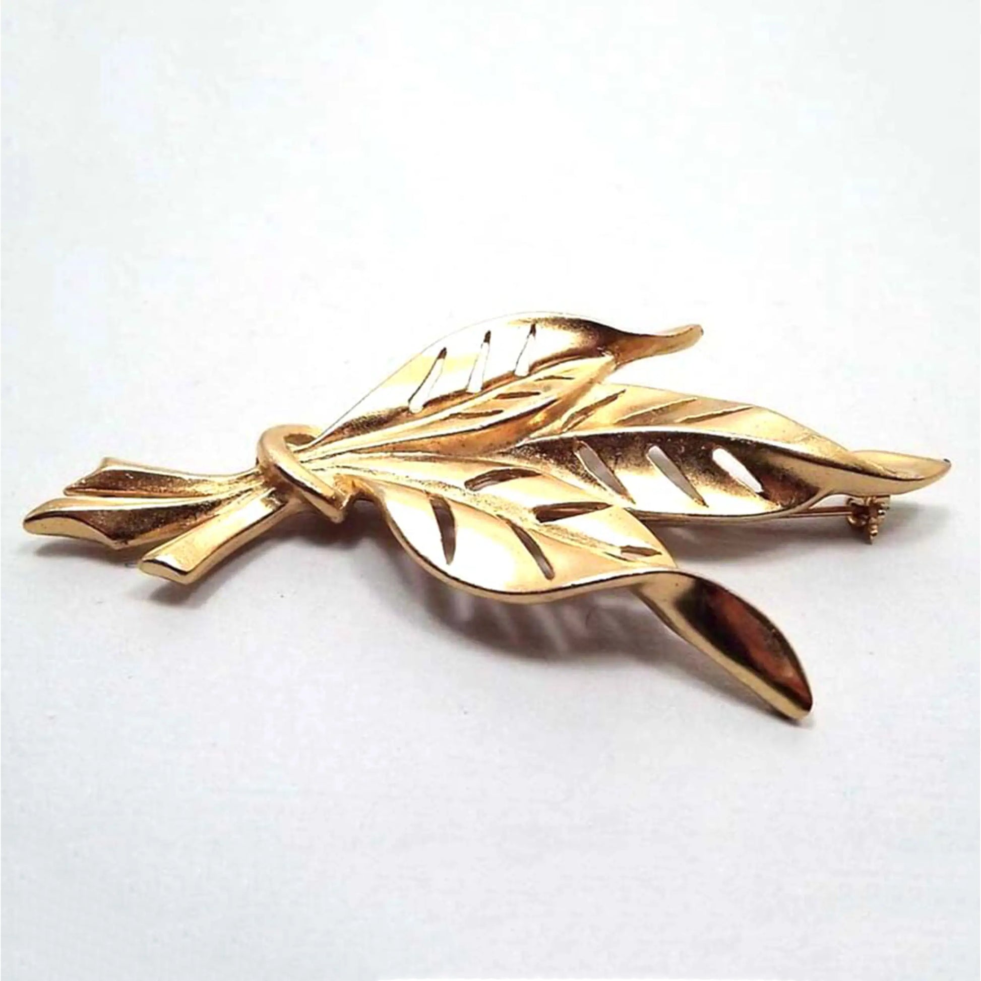 Front view of the retro vintage leaf brooch pin. It is matte gold tone in color with three leaves that are curvy with pointed ends and cut out areas for the leaf veins. The bottom stems look like they're tied together to form a bunch.