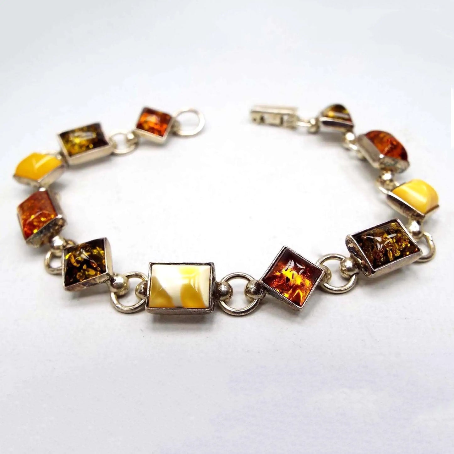 Top view of the Mid Century Baltic amber link bracelet. The setting is sterling silver and has alternating diamond shaped and rectangle shaped links. Each link has an amber cab. The amber has alternating colors of dark orange, olive green, and a yellow amber that is a marbled light to dark yellow color. There is a snap lock clasp on the end.