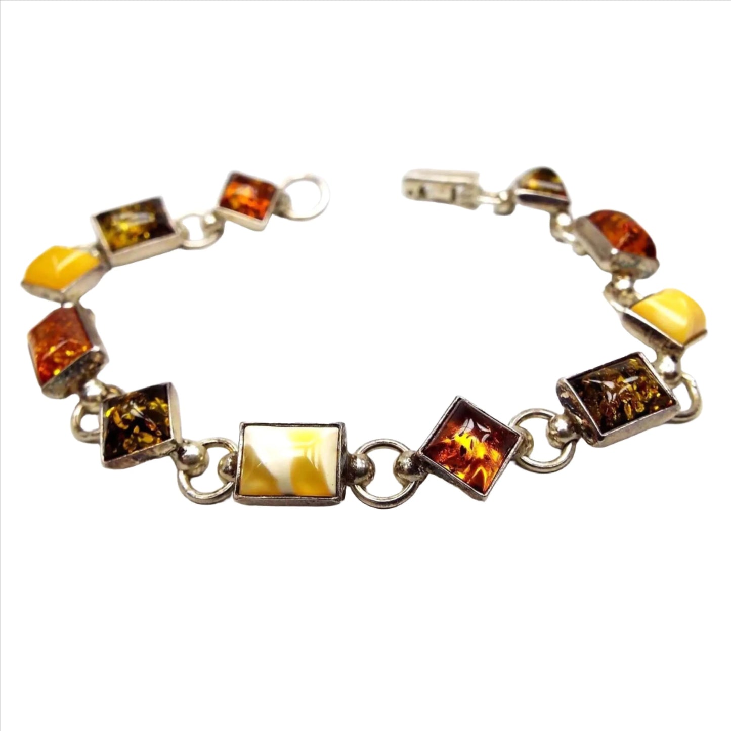 Top view of the Mid Century Baltic amber link bracelet. The setting is sterling silver and has alternating diamond shaped and rectangle shaped links. Each link has an amber cab. The amber has alternating colors of dark orange, olive green, and a yellow amber that is a marbled light to dark yellow color. There is a snap lock clasp on the end.