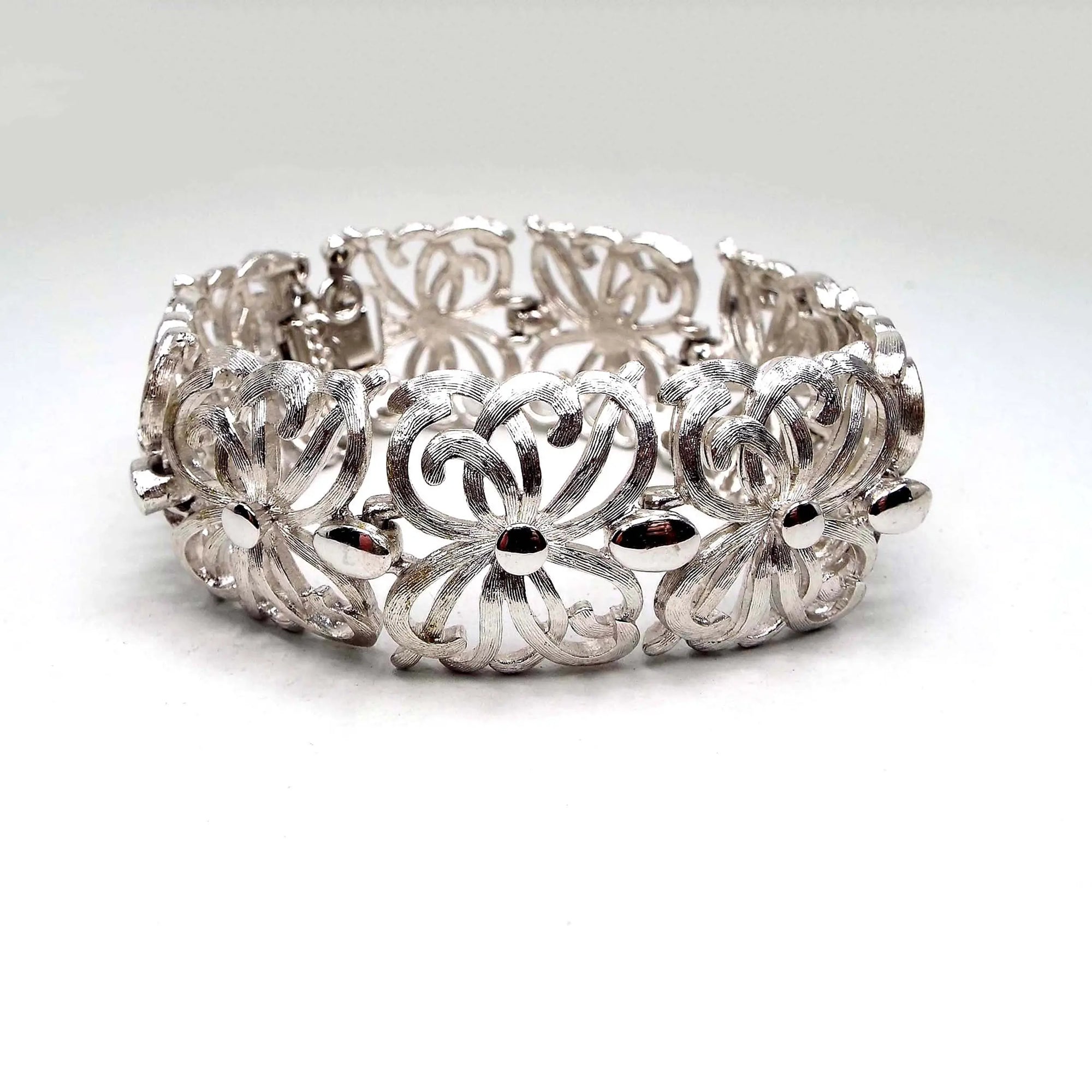 Angled front view of the Mid Century vintage Monet link bracelet. It is silver tone in color and has wide filigree links with a curvy floral like pattern. Most of the bracelet is textured matte metal. There is a snap lock clasp and safety chain on the end.