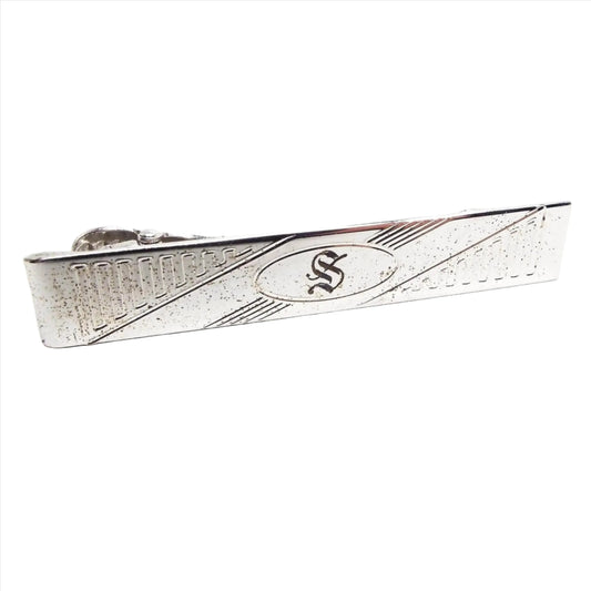 Front view of the Mid Century vintage Anson sterling silver initial tie clip. It is a long rectangle shape and the silver is slightly darkened with light pitting when seen under magnification. There is a rectangular line design on each side. The middle has an oval with the letter S engraved in it, and the oval has diagonal lines engraved on each side of the oval. 