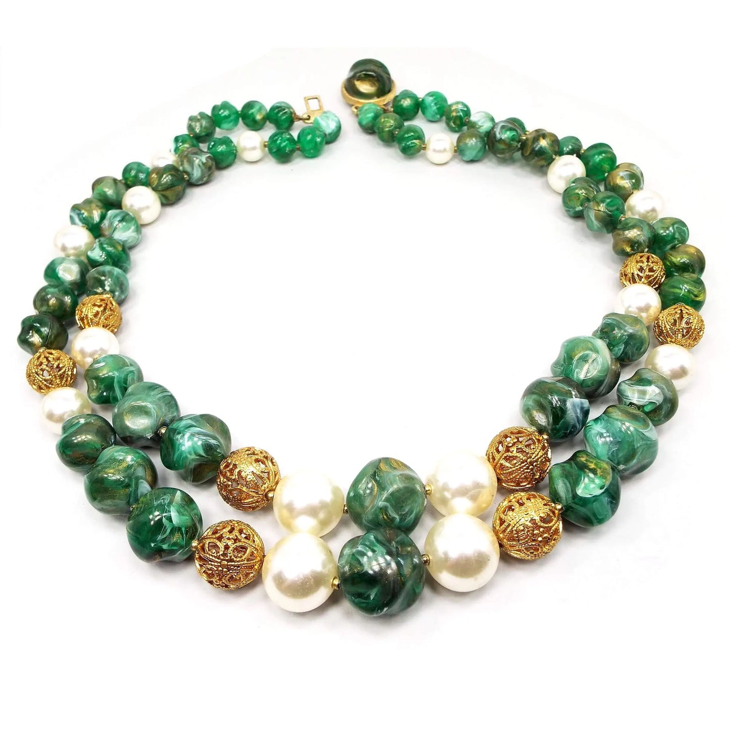 Angled view of the Mid Century vintage multi strand beaded necklace. There are two strands of chunky plastic beads that are shades of green with metallic gold swirled and marbled in. In between sets of those beads are plastic faux pearls and round gold tone plated filigree beads. There is a round clasp on the end with a domed cab that has the same coloring as the chunky beads. The metal components are gold tone plated in color.