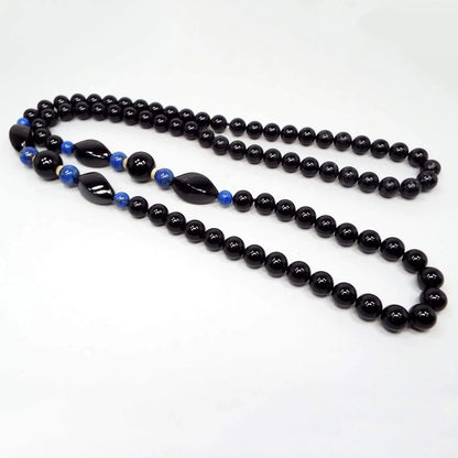 Top view of the retro vintage gemstone beaded necklace. The majority of the necklace is beaded with round ball black onyx beads. Partially the way up the necklace on each side is an area with smaller sized round ball blue lapis lazuli beads with twisted oval onyx beads and one larger onyx bead in the middle.
