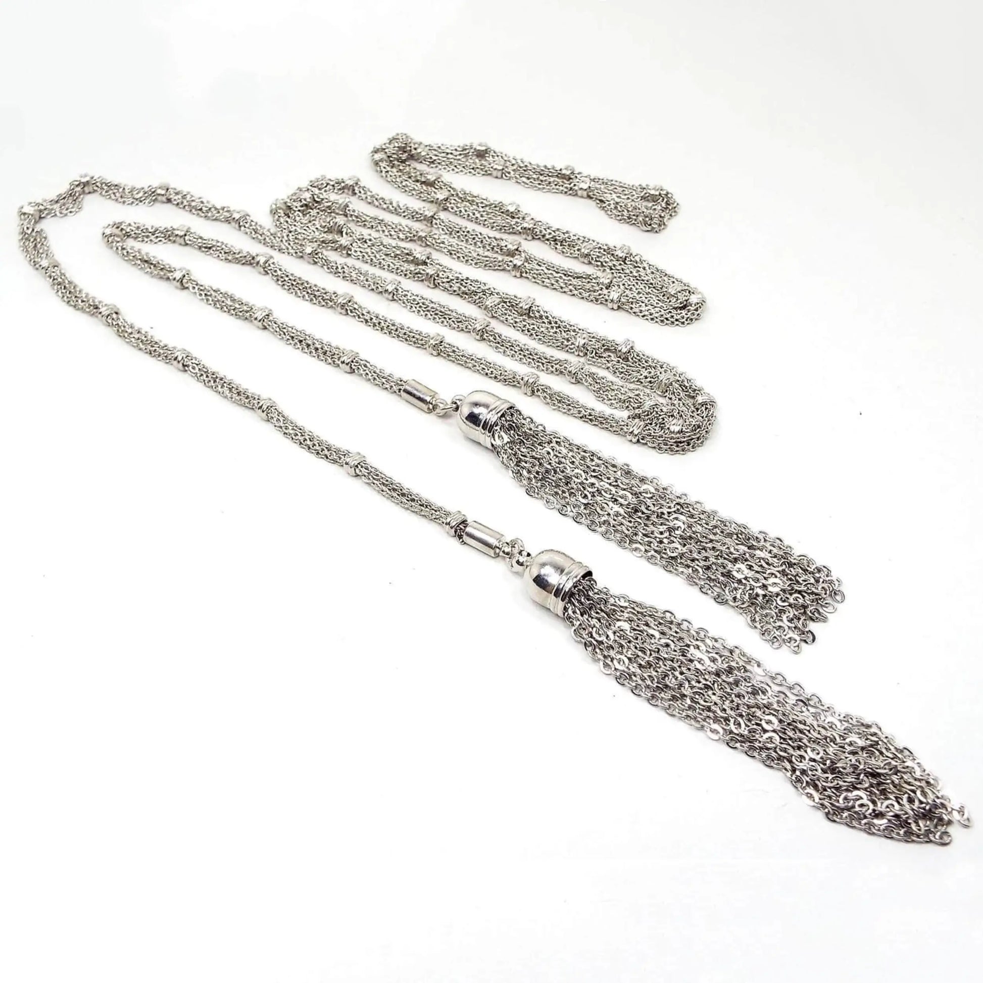 Angled top view of the long Mid Century vintage lariat chain tassel necklace. The metal is silver tone in color. the main part of the necklace has multi strands of oval link cable chain held together by spaced round open beads. The end tassels have a domed cap and several strands of round link rolo chain dangling from each end. There is no clasp as you wrap the necklace around yourself lariat style.