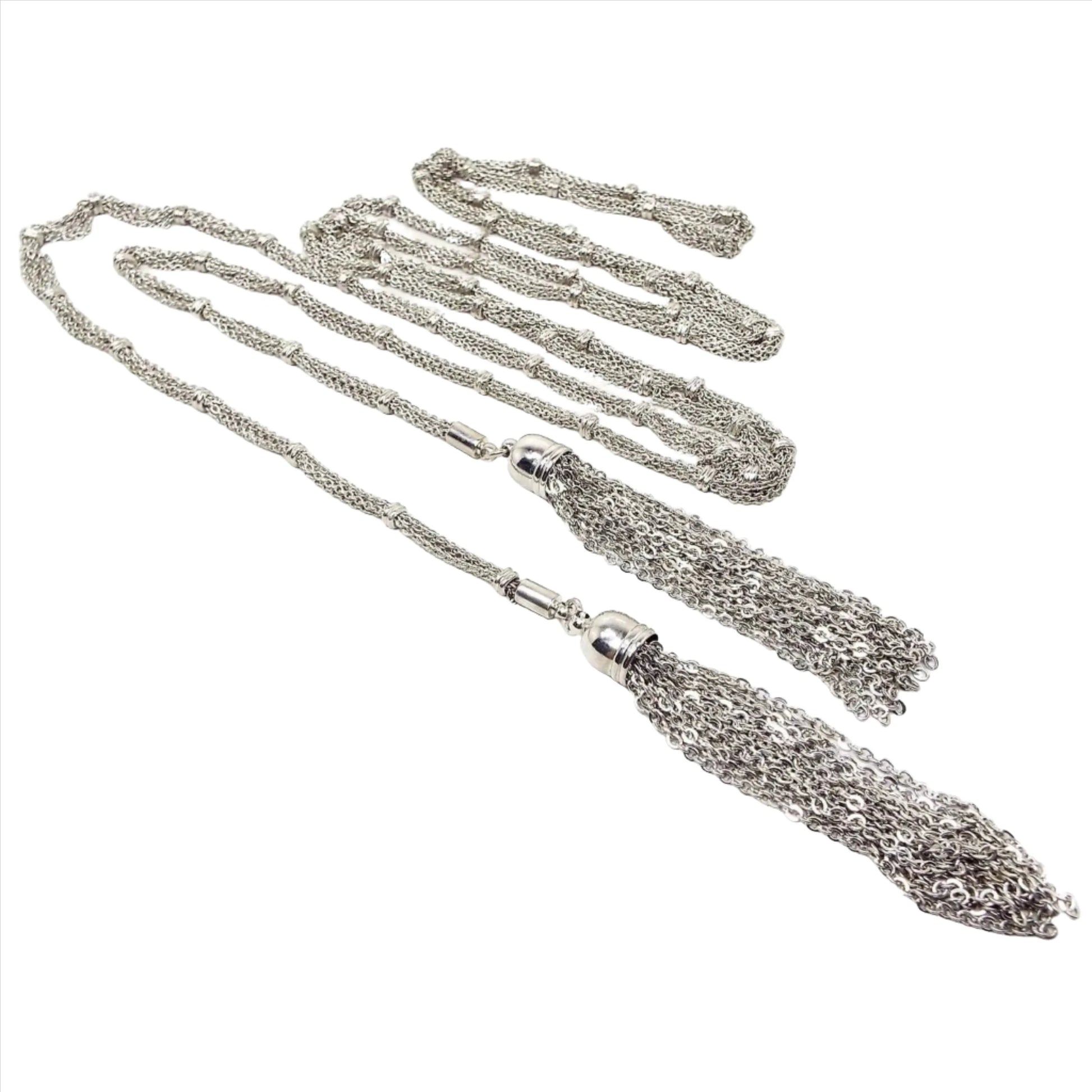 Angled top view of the long Mid Century vintage lariat chain tassel necklace. The metal is silver tone in color. the main part of the necklace has multi strands of oval link cable chain held together by spaced round open beads. The end tassels have a domed cap and several strands of round link rolo chain dangling from each end. There is no clasp as you wrap the necklace around yourself lariat style.
