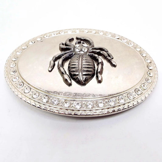 Front view of the retro vintage rhinestone goth spider belt buckle. The buckle is a large oval shape with a row of rhinestones around the edge. The very edge is lightly textured with a curved shape design. In the middle is a a raised tarantula with four rhinestones on its head. The spider is pewter gray in color and the rest of the belt buckle is shiny silver in color.