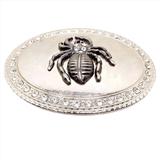 Front view of the retro vintage rhinestone goth spider belt buckle. The buckle is a large oval shape with a row of rhinestones around the edge. The very edge is lightly textured with a curved shape design. In the middle is a a raised tarantula with four rhinestones on its head. The spider is pewter gray in color and the rest of the belt buckle is shiny silver in color.