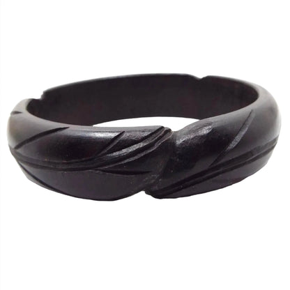 Side view of the retro vintage bangle bracelet. The bracelet is black painted wood with a basic carved feather design. There are two rounded feather shape ends slightly overlapping each other at the front of the bangle.It is a wider style bangle with a smooth inside area.